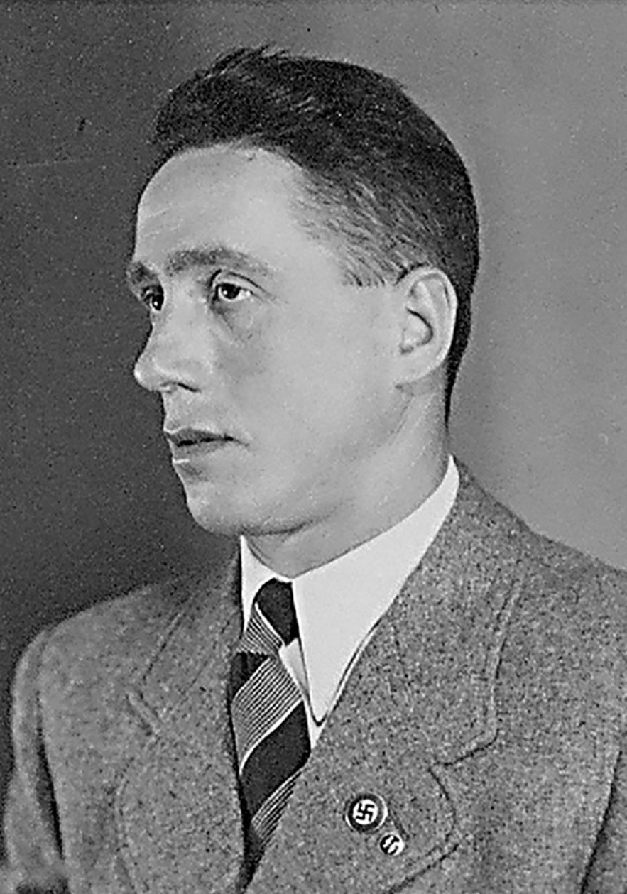 Halbprofil von Mann in hellem Anzug, wei&szlig;em Hemd und breitgestreifter Krawatte, schaut links an der Kamera weit vorbei. Hakenkreuz- und SS-Pinnadel am Revers. &copy; Deutsches Bundesarchiv