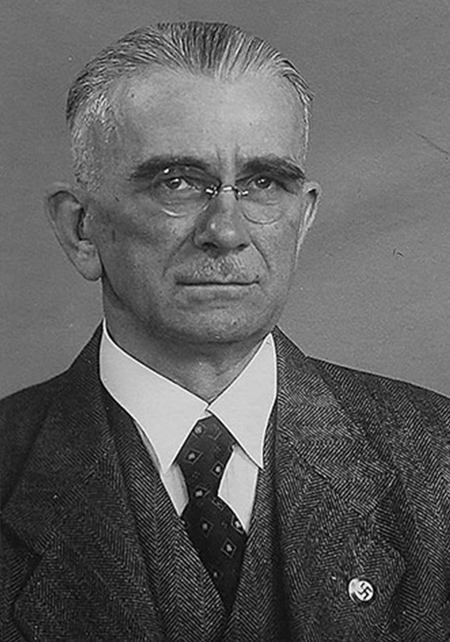 Frontal-Portrait von &auml;lterem Mann. Randlose Brille, eventuell ohne B&uuml;gel, Fischgr&auml;tenanzug und -Gilet, wei&szlig;es Hemd, gemusterte Krawatte, Hakenkreuz-Pinnadel am Revers. &copy; Wiener Stadt- und Landesarchiv