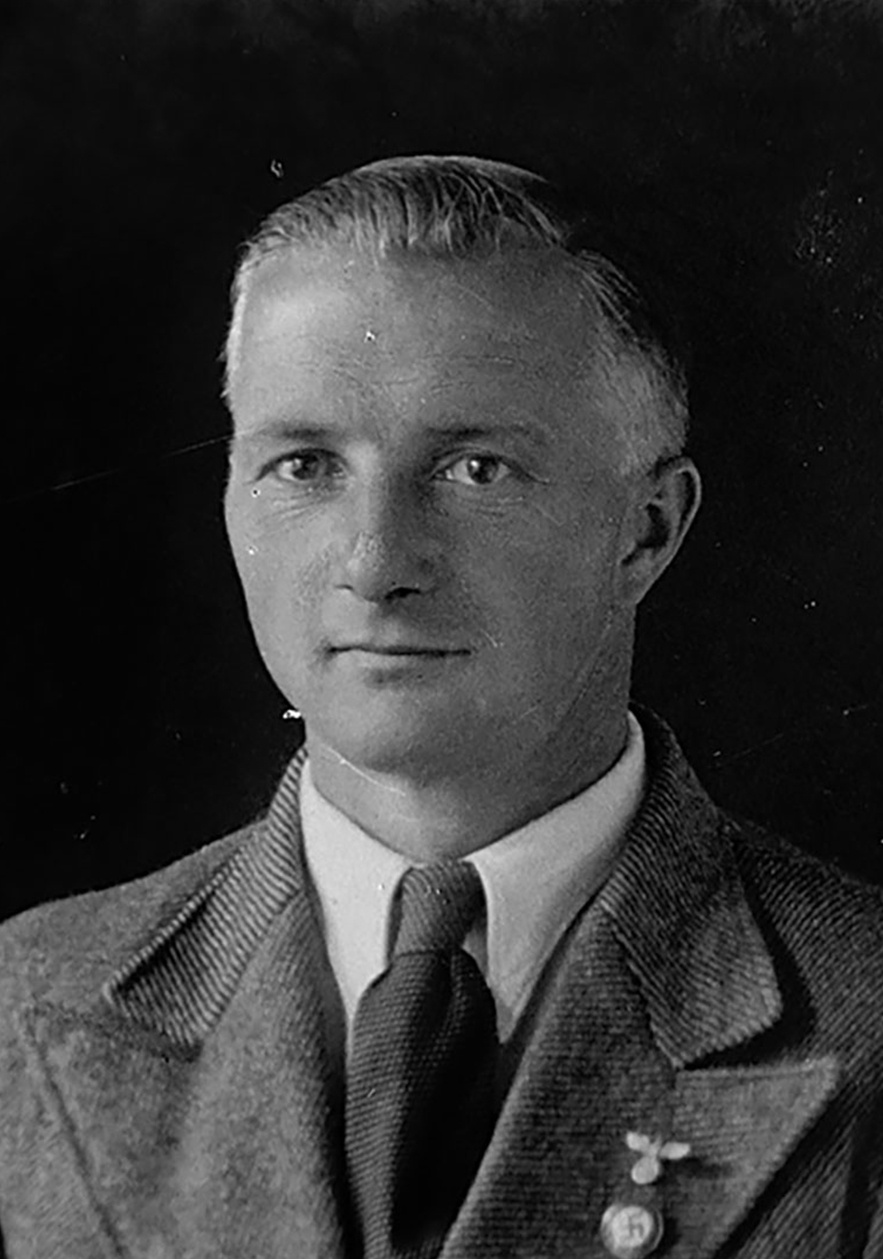 Frontalportrait eines Mannes mit kurzen vermutlich grauen Haaren, schmalgestreifter Anzug, wei&szlig;em Hemd, dunkler Krawatte, Hakenkreuz-Pinnadel am Revers. Blickt leicht links an der Kamera vorbei. &copy; Deutsches Bundesarchiv