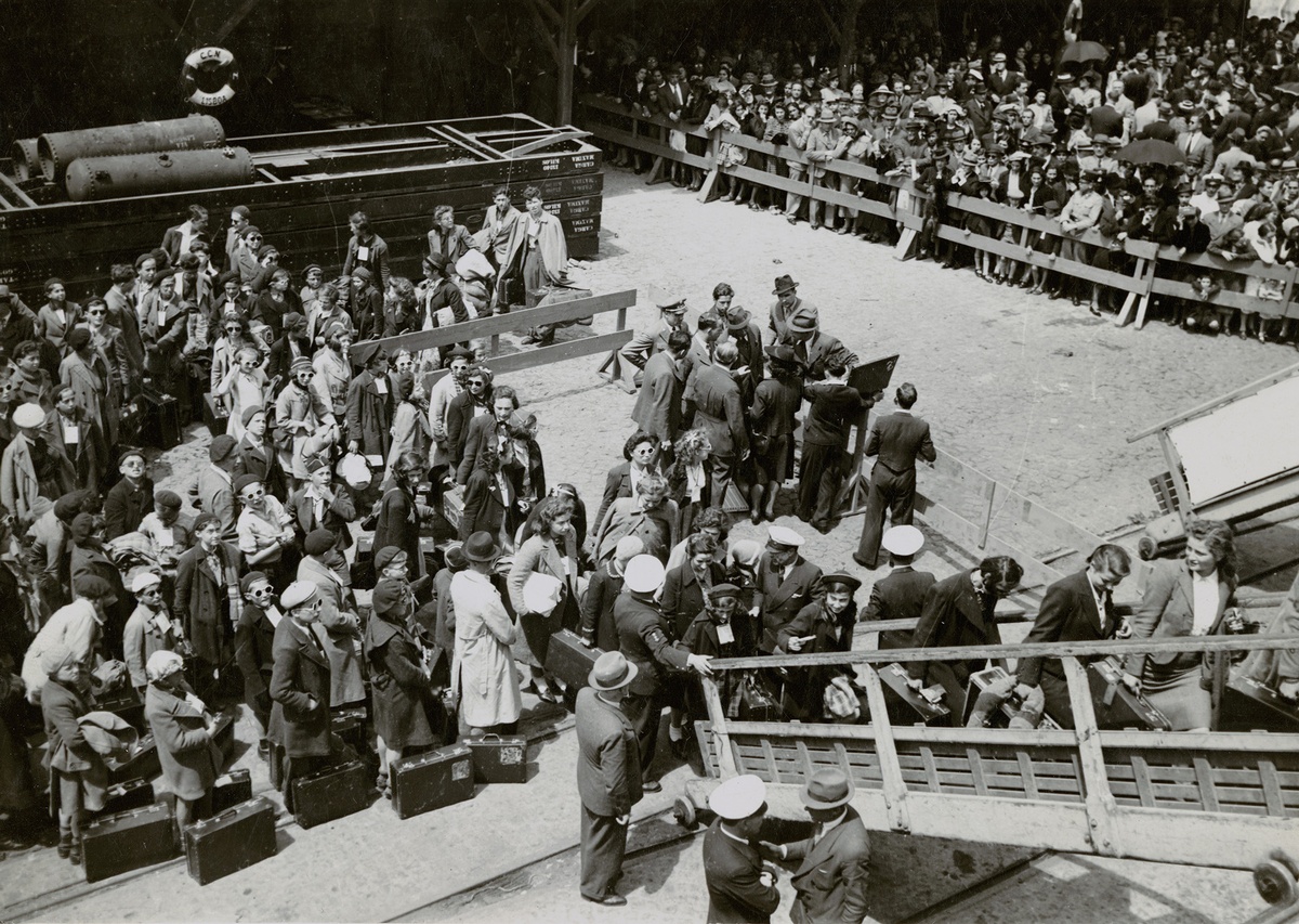 Menschen mit Gep&auml;ckst&uuml;cken stellen sich vor einer Treppe an. Links im Hintergrund ein Schwimmreifen auf einer Wand, rechts eine wartende Menschenmenge hinter einem Zaun. &copy; American Jewish Joint Distribution Committee Archives