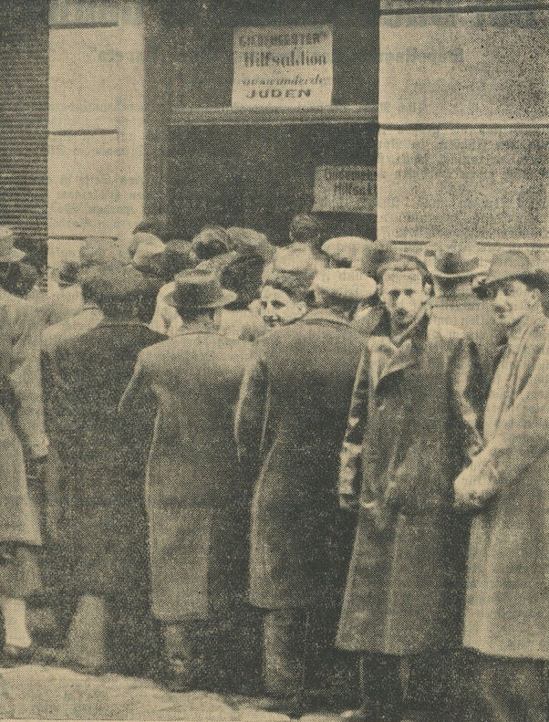 M&auml;nner mit M&auml;nteln und teils mit H&uuml;ten stehen Schlange vor einem Geb&auml;udeeingang, &uuml;ber dem ein Schild mit der Aufschrift &bdquo;Gildemeester Hilfsaktion&ldquo; befestigt ist. &copy; ANNO &Ouml;NB (V&ouml;lkischer Beobachter, Wiener Ausgabe, 13.5.1938)