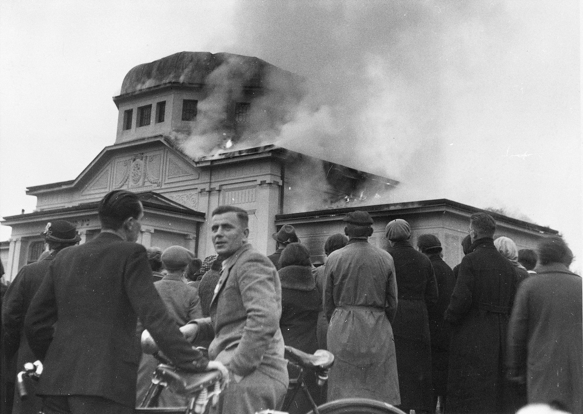Eine Menschenmenge blickt auf eine brennende Synagoge, aus der Rauchschwaden aufsteigen. Im Vordergrund ein Mann mit Fahrrad, der in Richtung Kamera blickt.  &copy; Universalmuseum Joanneum Graz/ Multimediale Sammlungen