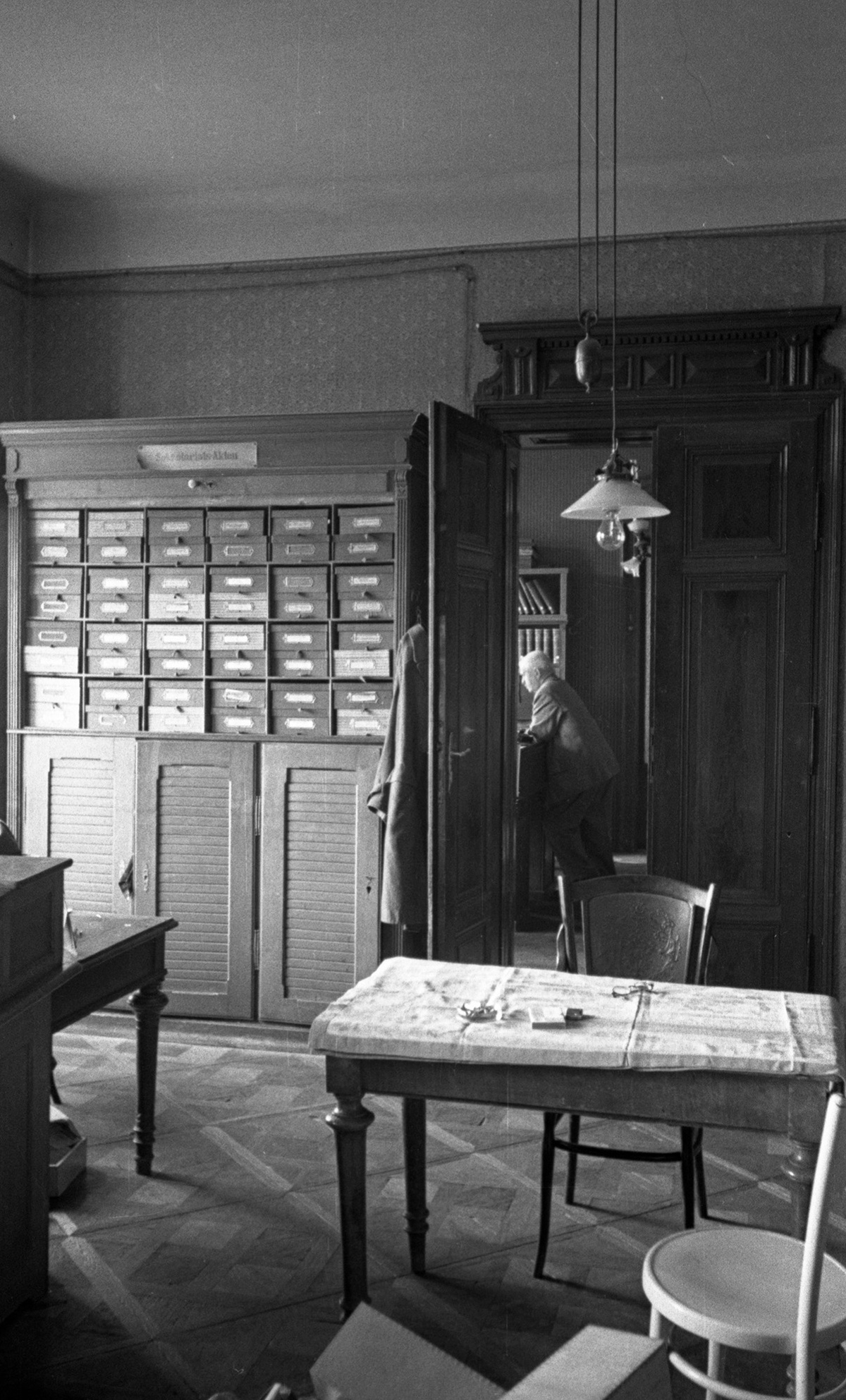 Tisch mit St&uuml;hlen im Vordergrund, links hinten an der Wand ein gro&szlig;er Aktenschrank, rechts daneben ein Durchgang in das Nebenzimmer, wo eine Person steht. &copy; Bundesarchiv