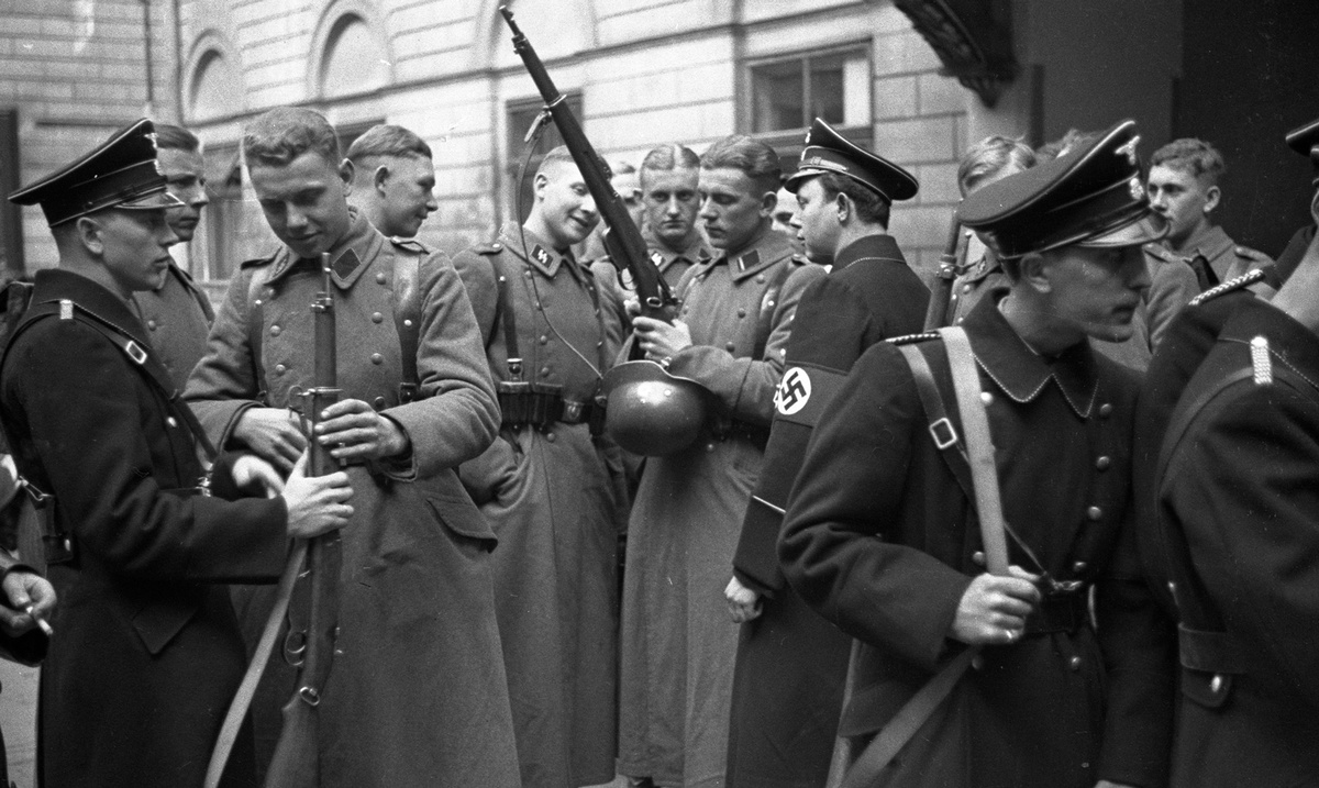 M&auml;nner in Uniform, teilweise mit Stahlhelm und Gewehr, stehen vor einem Geb&auml;ude in Gruppen zusammen. &copy; Bundesarchiv