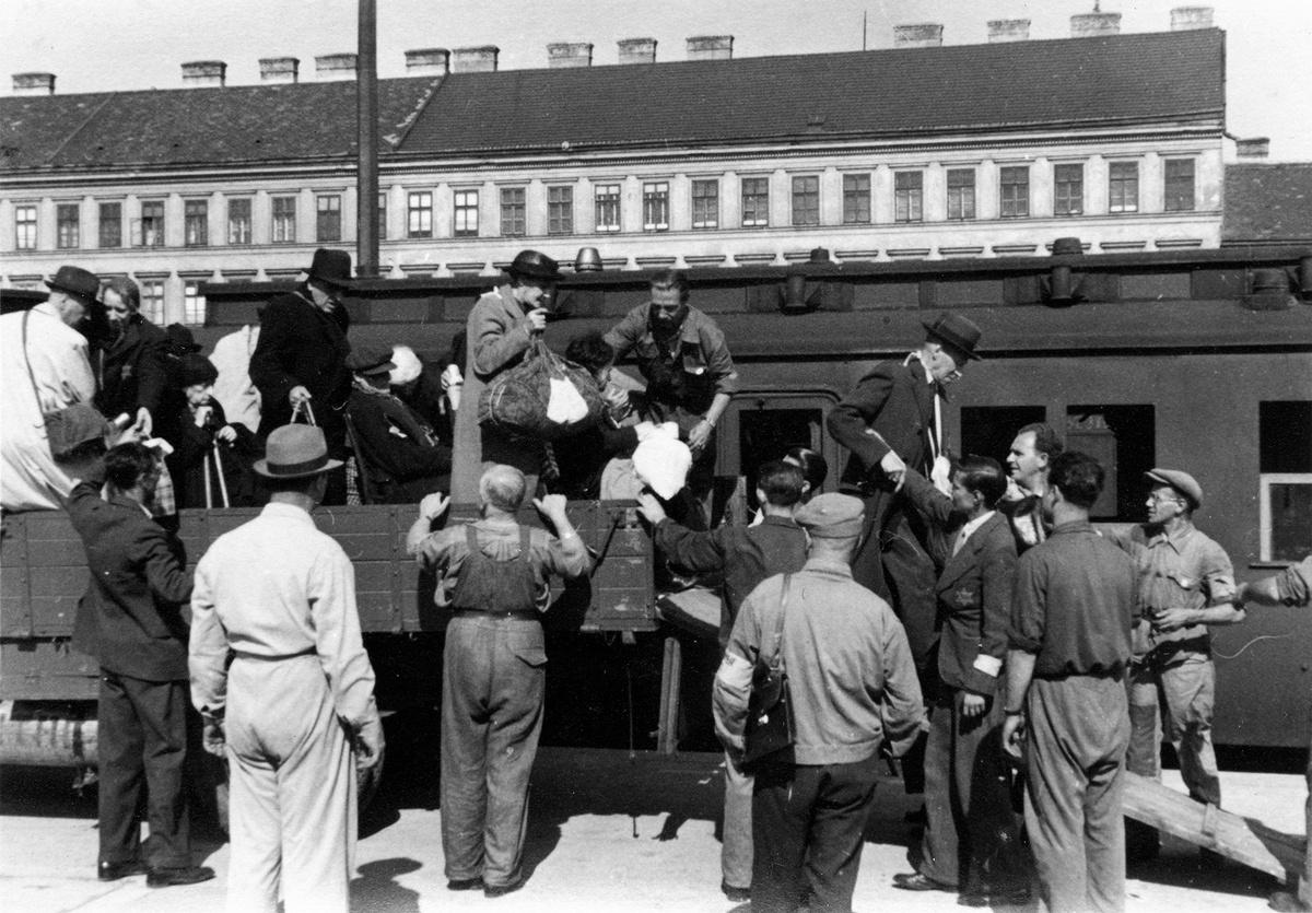 Mehrere Menschen stehen auf einem Anh&auml;nger und tragen Gep&auml;ckst&uuml;cke. M&auml;nner, zum Teil mit Armbinden und aufgen&auml;htem Stern, helfen ihnen beim Absteigen. Im Hintergrund ein Waggon, dahinter ein Geb&auml;ude. &copy; Bundesarchiv
