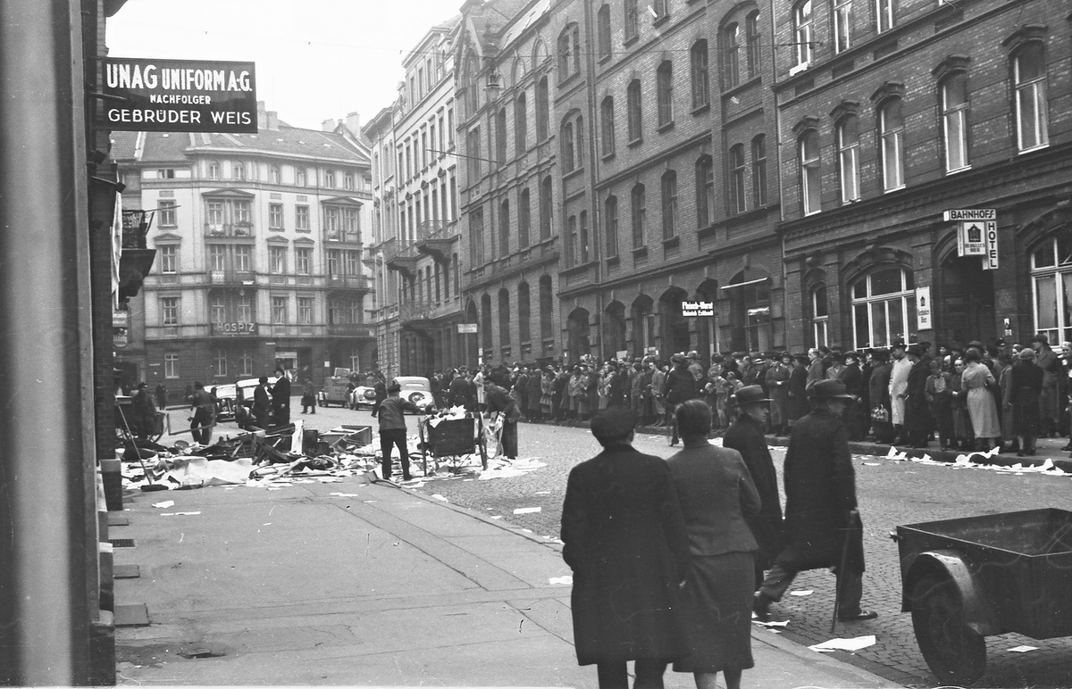 Eine Menschenmenge auf einer von H&auml;usern ges&auml;umten Stra&szlig;e, auf der zertr&uuml;mmerte Auslagenscheiben und demolierte M&ouml;bel liegen.  &copy; Stadtarchiv Kassel 