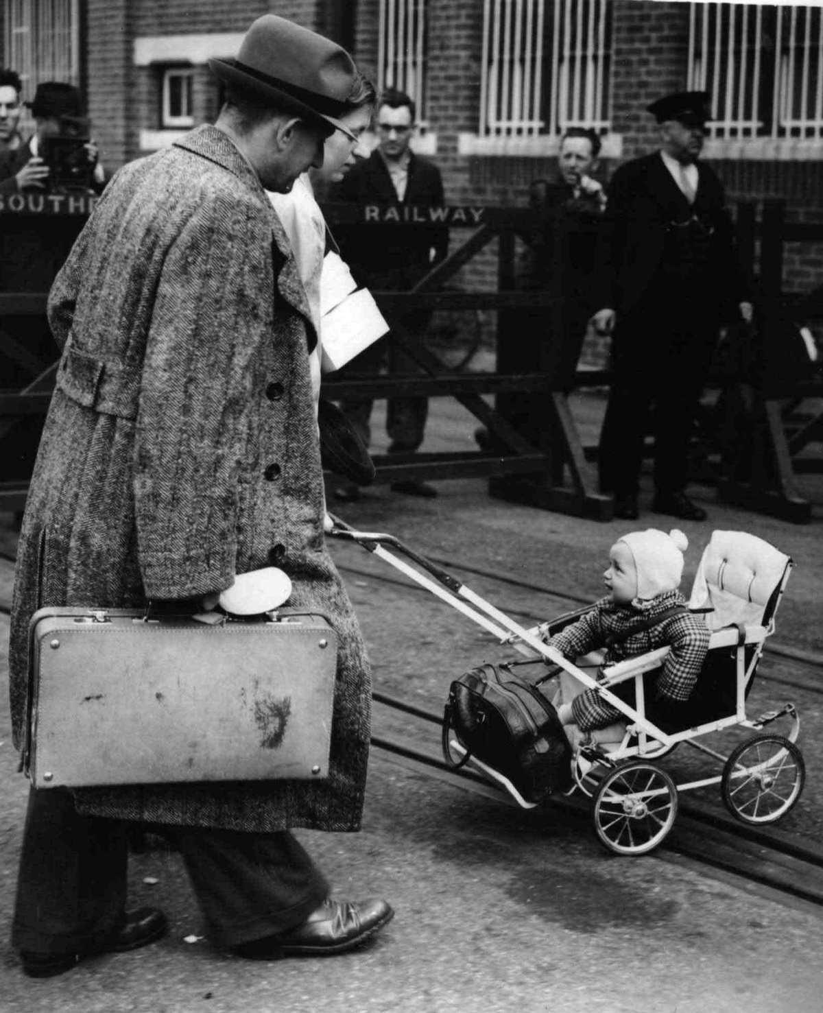 Mann mit einem Koffer in der Hand, langem Mantel und Hut geht neben einer Frau, die ein Kind mit Haube und Jacke in einem Kinderwagen &uuml;ber die Stra&szlig;e schiebt. Im Hintergrund mehrere Personen hinter einem Zaun, davor ein Mann mit Schaffnerkappe und Anzug.  &copy; &Ouml;NB Bildarchiv