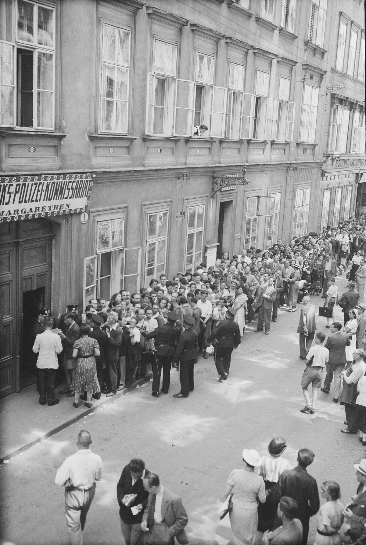 Eine lange Menschenschlange am Gehsteig vor einem Hauseingang, auf der Stra&szlig;e weitere Menschen. Ein Schild &uuml;ber dem Eingang mit der Aufschrift &bdquo;Polizeikommissariat Margareten&ldquo;, umrandet von kleinen Hakenkreuz-Symbolen. &copy; &Ouml;NB Bildarchiv