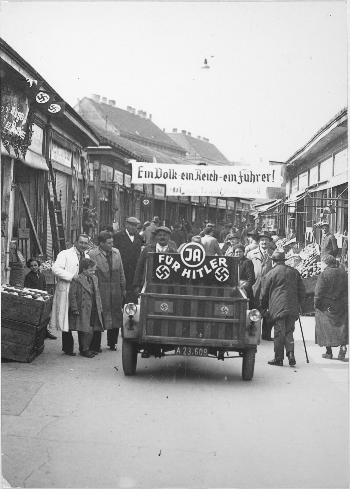 Eine von Marktst&auml;nden ges&auml;umte Stra&szlig;e, auf der ein Auto mit der Aufschrift &bdquo;Ja f&uuml;r Hitler&ldquo; und zwei Hakenkreuzen f&auml;hrt. Dahinter eine Menschenmenge. &Uuml;ber die Stra&szlig;e ist ein Transparent gespannt. Es tr&auml;gt die Aufschrift &bdquo;Ein Volk ein Reich ein F&uuml;hrer!&ldquo; &copy; &Ouml;NB Bildarchiv