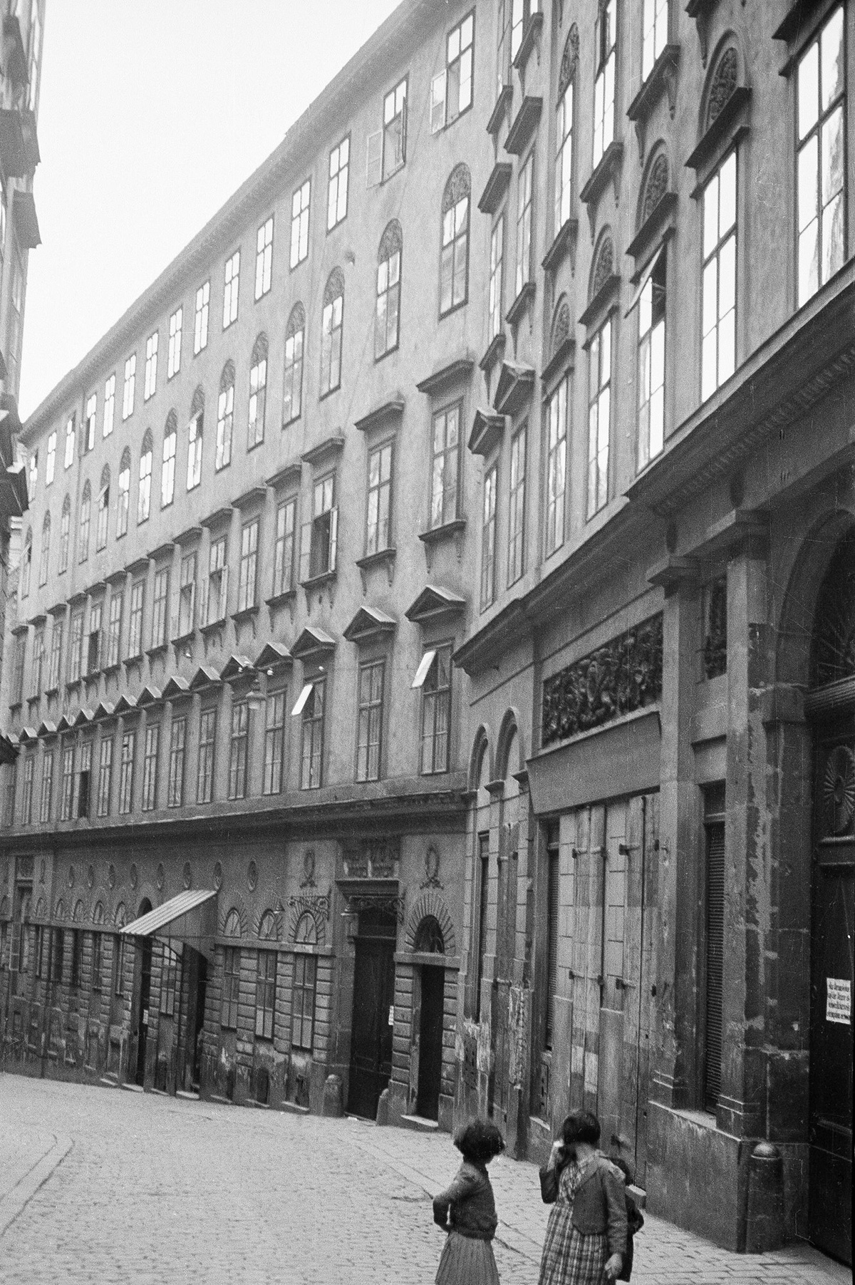 Stra&szlig;e mit H&auml;userfront, im Vordergrund zwei M&auml;dchen mit abgewandtem Blick.  &copy; &Ouml;NB Bildarchiv