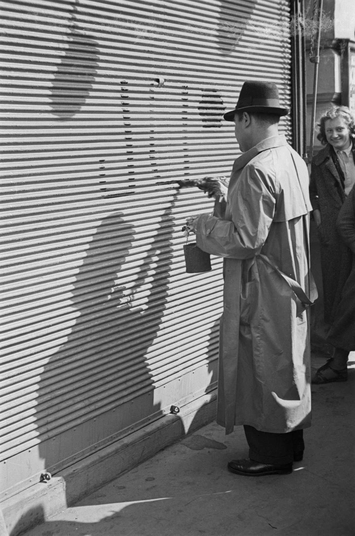 Ein Mann mit Hut und Mantel pinselt &bdquo;JU&ldquo; auf einen Rollladen. Eine lachende Frau sieht ihm zu. &copy; &Ouml;NB Bildarchiv 