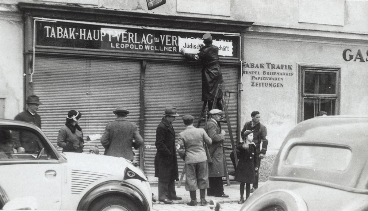 Gesch&auml;ftslokal mit der Aufschrift &bdquo;Tabak-Hauptverlag&ldquo;. Davor eine Gruppe von neun Personen und zwei Autos. Auf einer Leiter steht ein Mann, der &uuml;ber dem Eingang des Gesch&auml;fts eine Tafel mit der Aufschrift &bdquo;J&uuml;disches Gesch&auml;ft&ldquo; befestigt.  &copy; &Ouml;NB Bildarchiv
