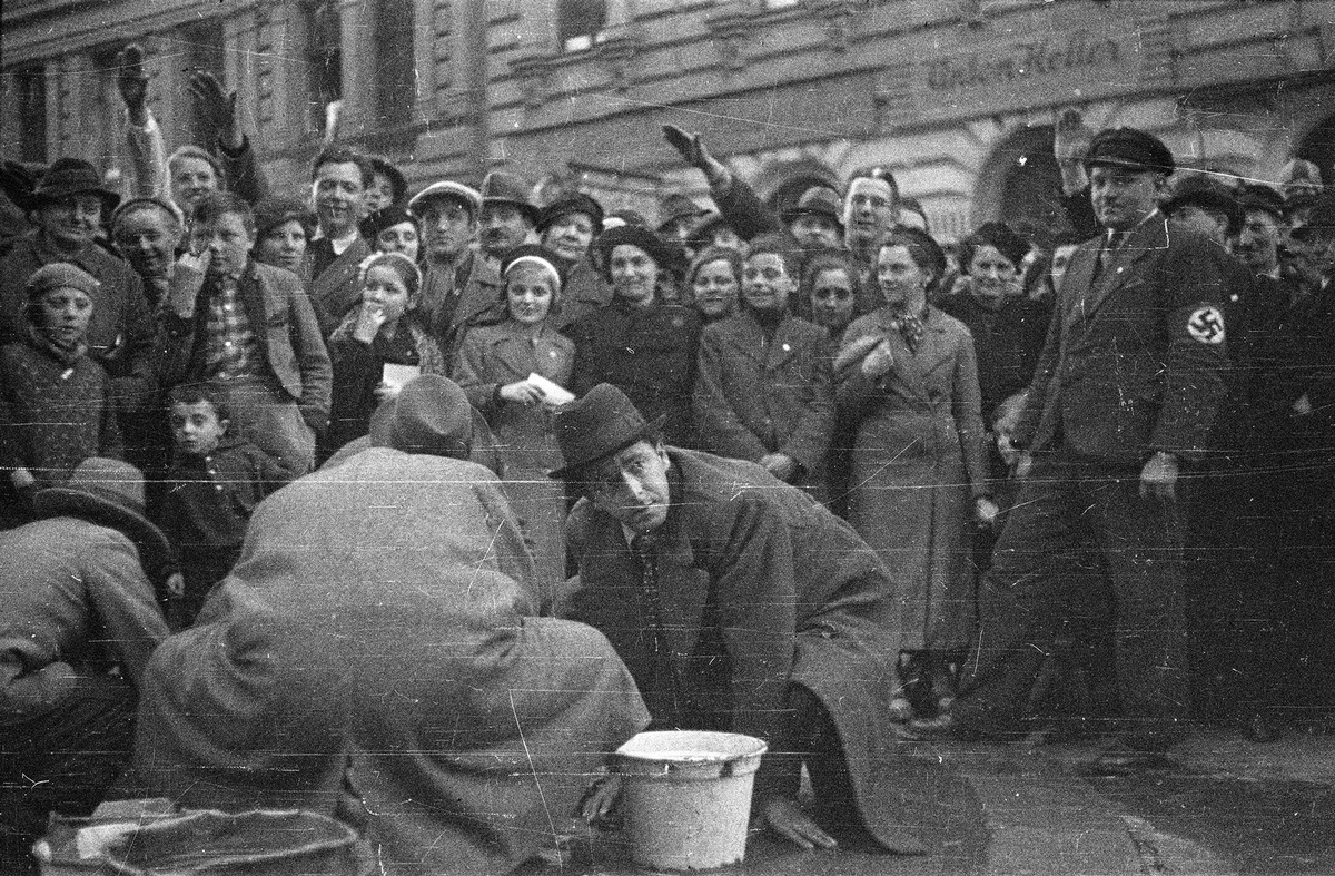 Eine Menschenmenge steht vor einem Geb&auml;ude, teils recken die Menschen die rechte Hand in die H&ouml;he, ein Mann im Vordergrund rechts tr&auml;gt eine Hakenkreuzbinde am Arm. Im Zentrum des Bildes knien drei M&auml;nner am Boden, neben ihnen stehen Wassereimer. &copy; &Ouml;NB Bildarchiv 
