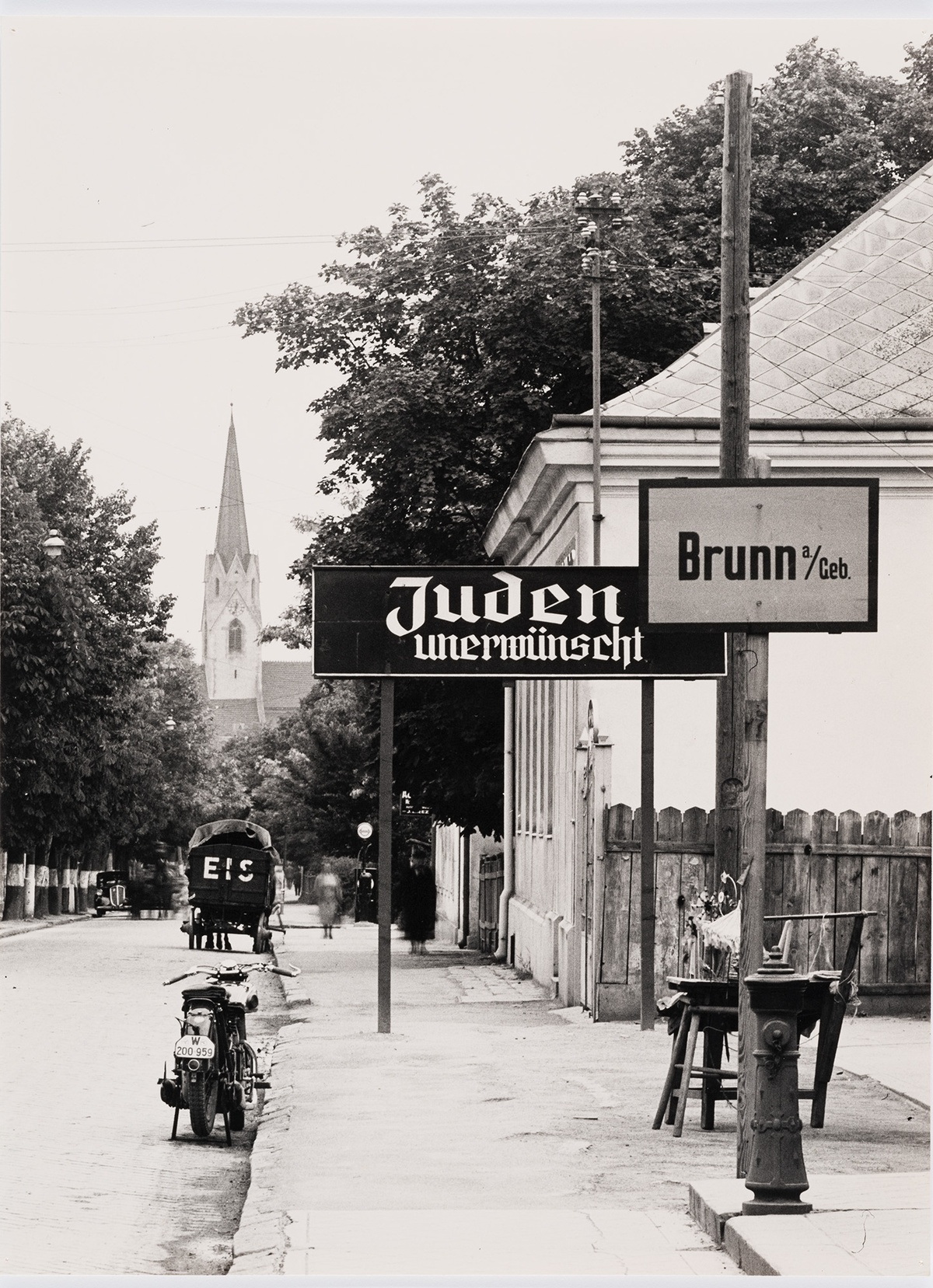 Ortseingang von Brunn am Gebirge. Hinter der Ortstafel steht ein Schild mit der Aufschrift &bdquo;Juden unerw&uuml;nscht&ldquo;. Im Hintergrund ein Haus und eine Kirche. &copy; Wien Museum