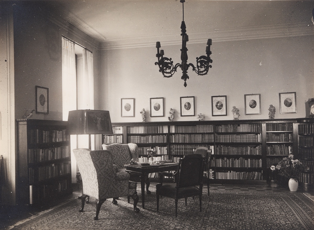 immer mit B&uuml;cherwand, dar&uuml;ber eine Reihe von gerahmten Bildern an der Wand und ein Kronleuchter. Davor ein Tisch mit gepolsterten St&uuml;hlen auf einem gro&szlig;en Teppich. Rechts im Eck eine Vase. &copy; Wien Museum 