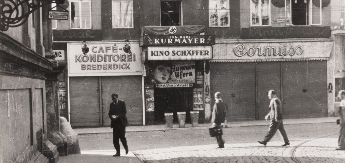 Fassade eines Hauses, mittig ein Hauseingang, dar&uuml;ber eine Leuchtreklame &bdquo;Kino Sch&auml;ffer&ldquo;, darunter ein Schild mit der Aufschrift &bdquo;Arisierter Betrieb&ldquo;. Im Vordergrund zwei Passanten. &copy; Wien Museum