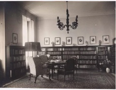 Zimmer mit B&uuml;cherwand, dar&uuml;ber eine Reihe von gerahmten Bildern an der Wand und ein Kronleuchter. Davor ein Tisch mit gepolsterten St&uuml;hlen auf einem gro&szlig;en Teppich. Rechts im Eck eine Vase. &copy; Wien Museum 